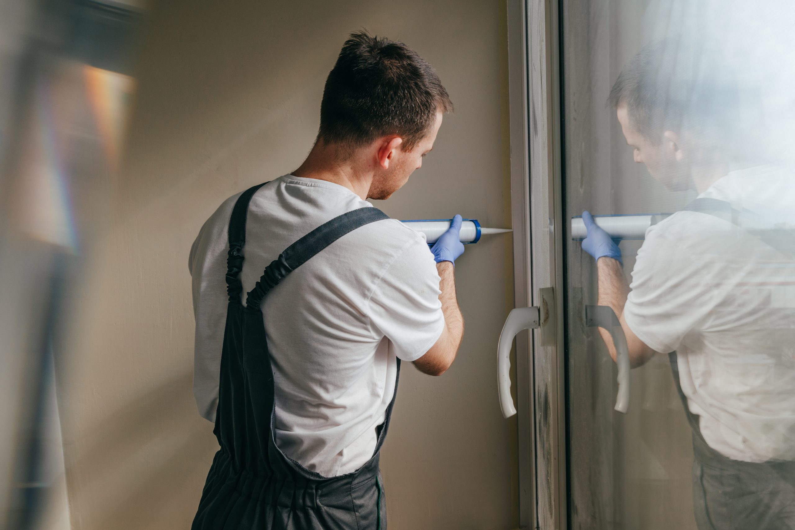 Young man wearing overalls sealing cracks between window and trim using waterproof silicone caulk on the balcony.